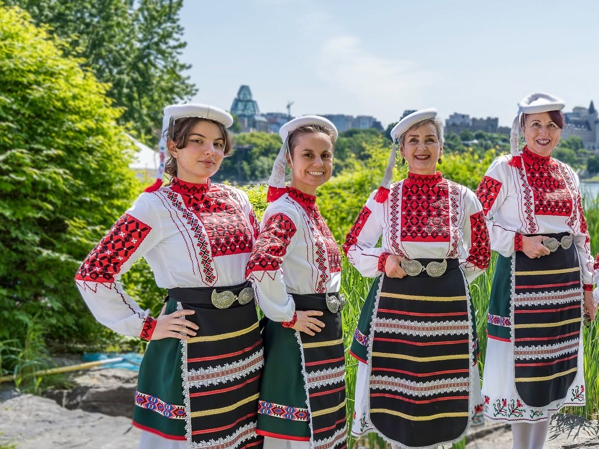 Four Rodina dancers in Bulgarian folk costumes beside the Ottawa River, with Parliament in the background