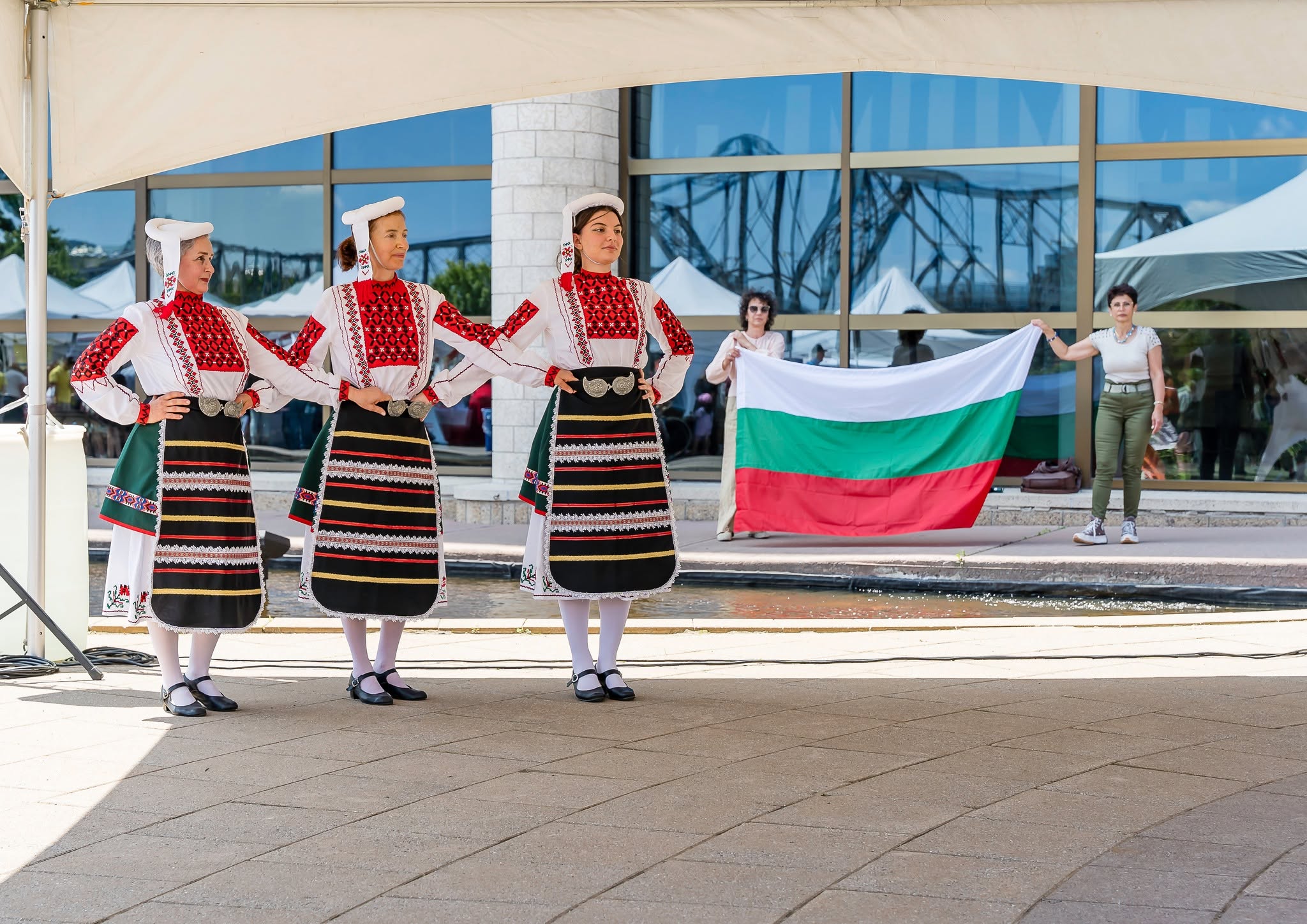Three Rodina dancers performing in Bulgarian folk costumes with the Bulgarian flag behind the stage